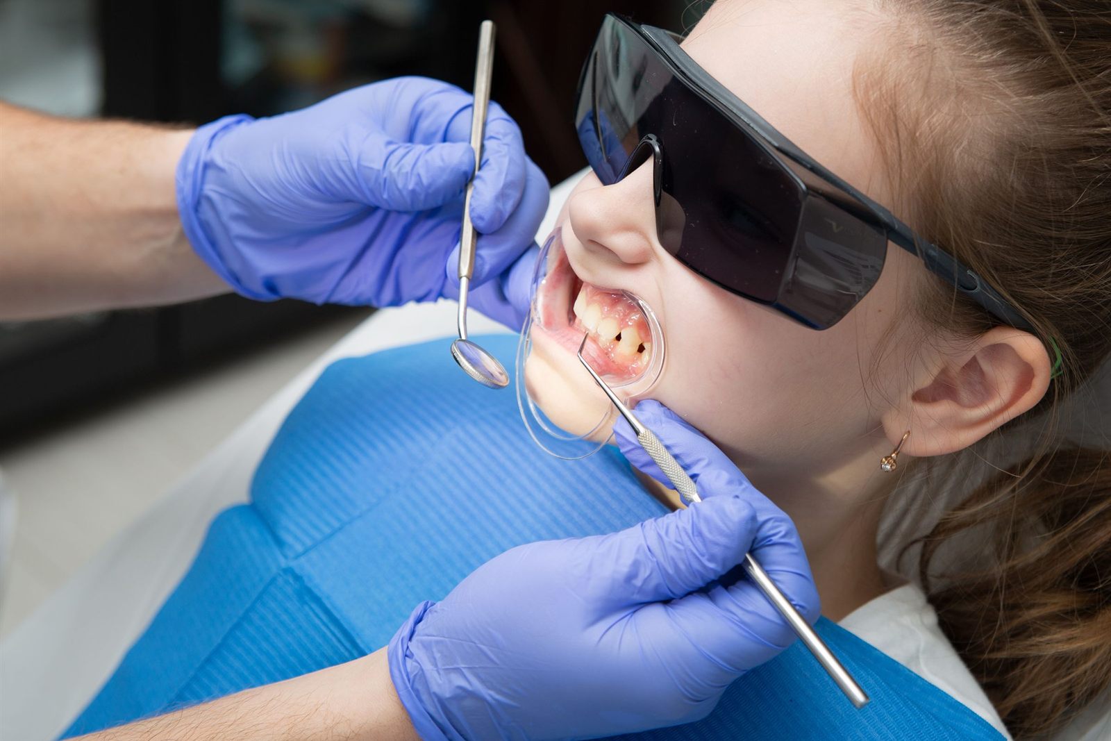 Dentist checking a young girl's back teeth with a dental mirror while she wears protective glasses