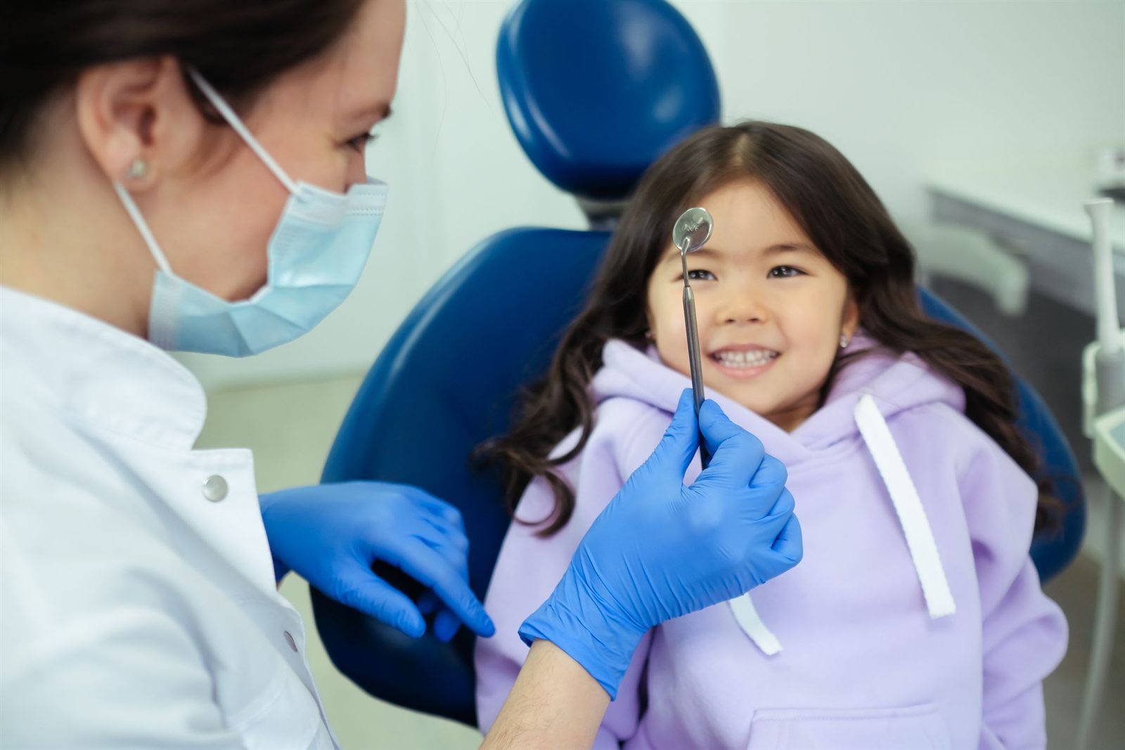Smiling girl in a dental chair while her dentist holds up a dental mirror
